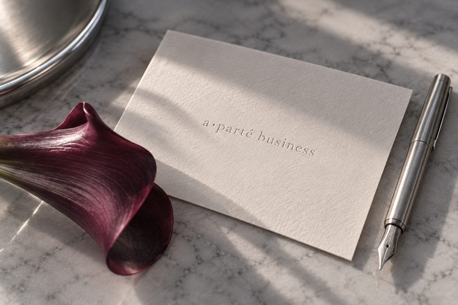 An embossed a•parté business card on marble beside a burgundy calla lily and a silver pen